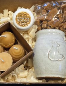 Gift box arrangement featuring a round jar labeled Natural Beeswax Moisturizing Skin Cream Lavender, beige soaps, and a ceramic vessel among wood shavings.